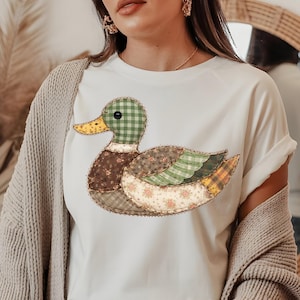 May include: White t-shirt featuring a patchwork duck design. The duck has a green checkered head, brown body, and wings with floral, plaid, and patterned fabrics. The duck's beak is yellow. The shirt has rolled-up sleeves.