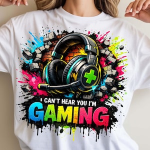 May include: White t-shirt featuring a graphic of a gaming headset with a green plus sign, set against a brick wall and colorful paint splatters. Text reads "I can't hear you I'm GAMING" in a vibrant, multi-colored font.