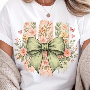 May include: White t-shirt with a light green bow illustration as the central design. The bow is set against vertical stripes with floral and botanical elements in soft pinks, oranges, and greens. Small pink hearts and butterflies complete the design.