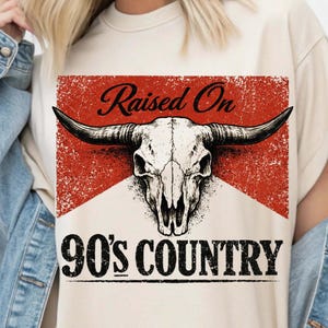 May include: Cream-colored t-shirt featuring a distressed red and white graphic with a bull skull and the text "Raised On 90's COUNTRY" in black. The shirt has a classic crew neck.