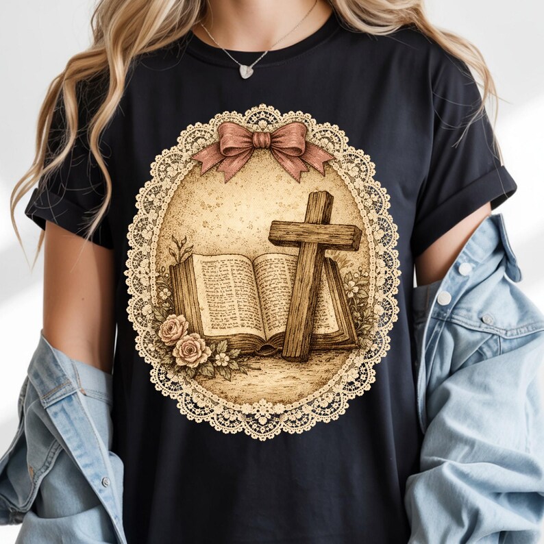 May include: Black t-shirt with a sepia-toned graphic of a wooden cross, open book, and roses inside a lace-edged oval. A pink bow is at the top. The design has a vintage, religious theme.
