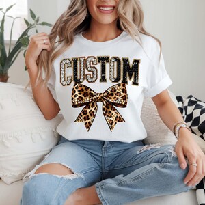 Pu&ograve; includere: T-shirt bianco con la scritta "CUSTOM" in strass e lettere nere sopra un fiocco leopardato. Il fiocco ha dettagli dorati e strass. La maglietta &egrave; di stile casual.