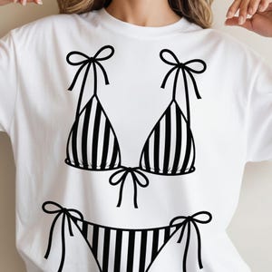 K&ouml;nnte beinhalten: Wei&szlig;es T-Shirt mit einer schwarz-wei&szlig;en Bikini-Grafik. Das Bikini-Oberteil und -Unterteil sind gestreift, mit Schleifen an den Tr&auml;gern und Seiten. Ein schlichtes, k&uuml;hnes Design, ideal f&uuml;r den Sommer.