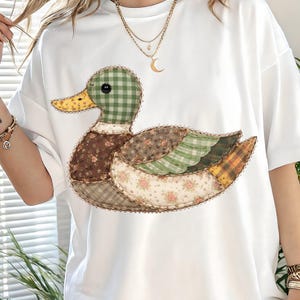 May include: White t-shirt featuring a patchwork duck design. The duck has a green checkered head, a yellow beak, and a body composed of various floral and plaid fabrics in shades of brown, green, and yellow. The shirt is a casual, oversized style.