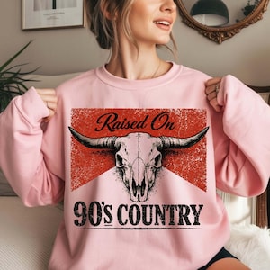 May include: Pink sweatshirt with a graphic design featuring a bull skull and the text "Raised On 90's Country". The design is set against a red, distressed background. The sweatshirt has long sleeves.