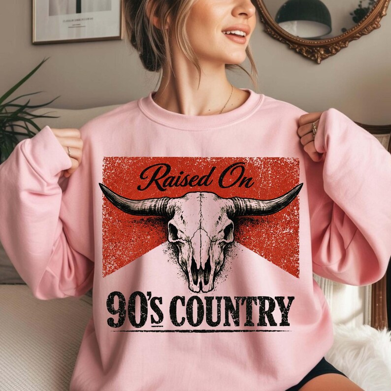 May include: Pink sweatshirt with a graphic design featuring a bull skull and the text "Raised On 90's Country". The design is set against a red, distressed background. The sweatshirt has long sleeves.