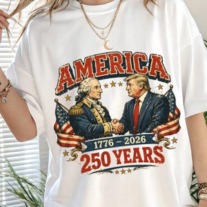 May include: White t-shirt featuring a graphic with the word "AMERICA" in red and blue, with an illustration of two men shaking hands, American flags, and the text "1776-2026 250 YEARS."