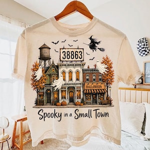 Op de afbeelding: Een crèmekleurig t-shirt met een afbeelding van een klein stadje met een watertoren, een reclamebord met het getal 38863, geesten, vleermuizen, een heks op een bezemsteel en pompoenen. De tekst luidt Spooky in a Small Town.