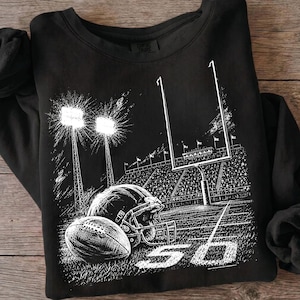 May include: A black long-sleeve sweatshirt featuring a white graphic print of a football stadium at night. The design includes a football, a helmet, goalposts, stadium lights, and the number 50 on the field, with a Comfort Colors brand label.