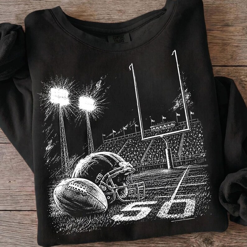 May include: A black long-sleeve sweatshirt featuring a white graphic print of a football stadium at night. The design includes a football, a helmet, goalposts, stadium lights, and the number 50 on the field, with a Comfort Colors brand label.