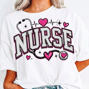 Puede incluir: Una camiseta blanca con la palabra NURSE en letras universitarias grandes, blancas con puntos y contorno negro. El dise&ntilde;o incluye un gr&aacute;fico de estetoscopio negro, varios corazones rosas brillantes y una l&iacute;nea de latido.