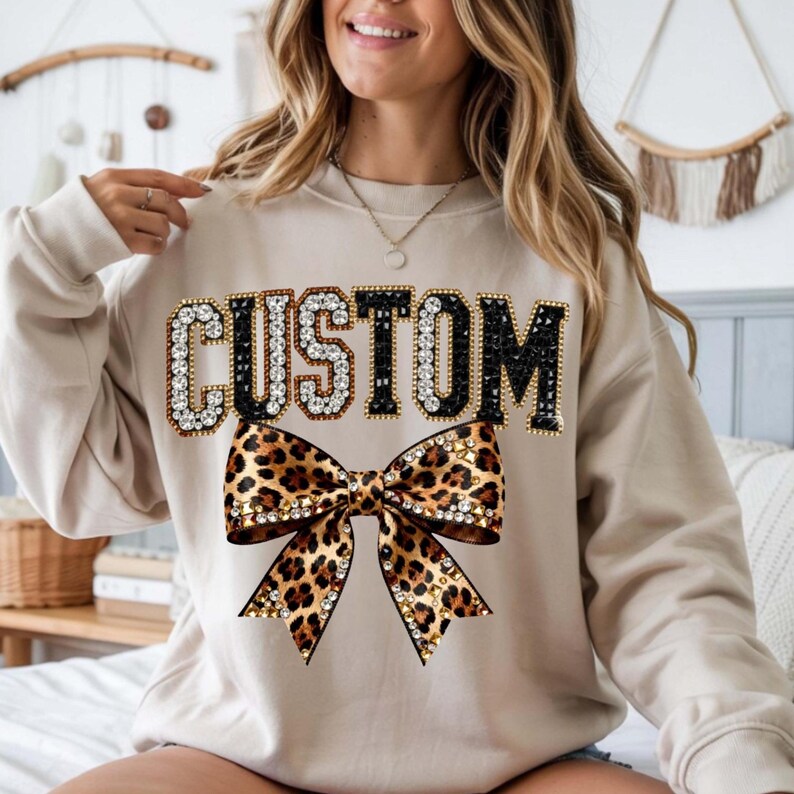 Pu&ograve; includere: Felpa beige con la scritta "CUSTOM" in un font gioiello. Sotto, un fiocco leopardato con dettagli di strass. La felpa &egrave; in stile casual.