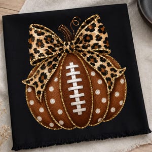 May include: A black fabric square featuring a central graphic of a brown pumpkin shaped like a football with white laces. The pumpkin is decorated with white polka dots and topped with a large leopard print bow with gold glitter trim.