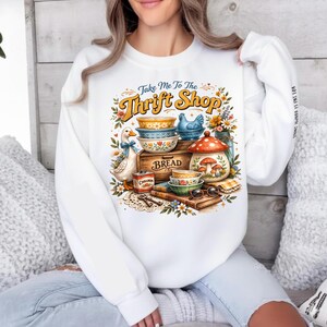 K&ouml;nnte beinhalten: Wei&szlig;es Sweatshirt mit einem farbenfrohen Aufdruck mit der Aufschrift "Take Me To The Thrift Shop". Das Design umfasst eine Ente mit blauer Schleife, Schalen, eine Brotkiste und einen Topf mit Pilzdekor. Der Text auf dem &Auml;rmel ist teilweise sichtbar.