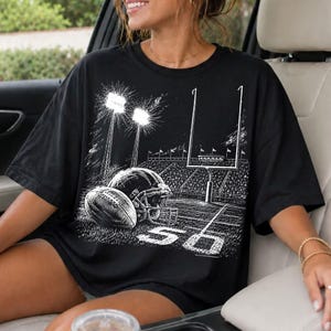 May include: A black short-sleeve t-shirt featuring a white graphic print of a football stadium at night. The design includes a football, a helmet, stadium lights, goalposts, and the number 50 marked on the field in a sketch-like style.