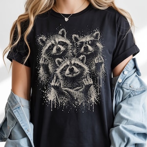 May include: Black t-shirt featuring a graphic of three raccoons giving the middle finger. One raccoon wears sunglasses. The design is in shades of gray and white, with a distressed, splattered effect.