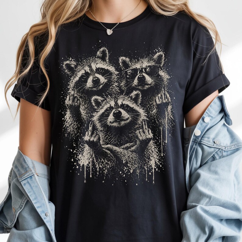 May include: Black t-shirt featuring a graphic of three raccoons giving the middle finger. One raccoon wears sunglasses. The design is in shades of gray and white, with a distressed, splattered effect.
