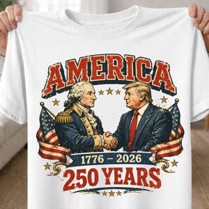 May include: White t-shirt featuring a graphic with the word "AMERICA" in red and blue. The graphic depicts two men shaking hands, with American flags and the text "1776-2026 250 YEARS".