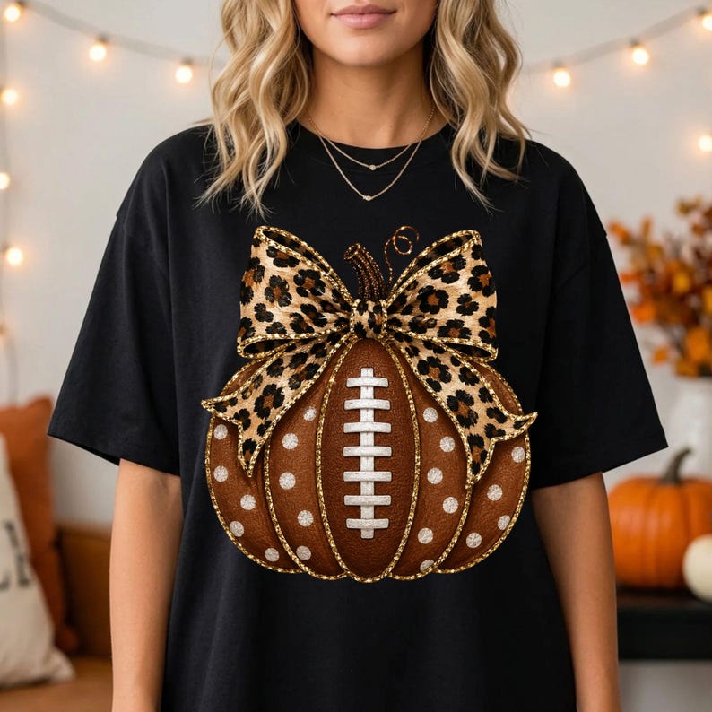 May include: A black t-shirt featuring a central graphic of a brown pumpkin with white polka dots and a football lace design in the center. The pumpkin is topped with a large, decorative bow featuring a brown and black leopard print pattern.