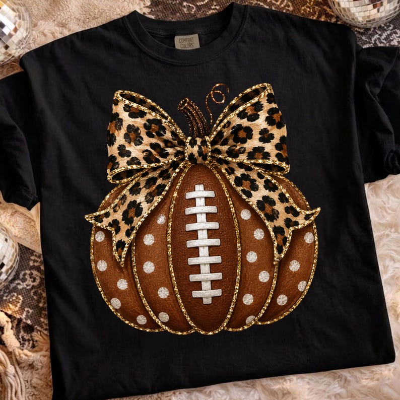 May include: A black t-shirt featuring a graphic of a brown pumpkin designed to look like a football with white laces. The pumpkin is decorated with white polka dots and topped with a large leopard print bow with gold glitter edges.