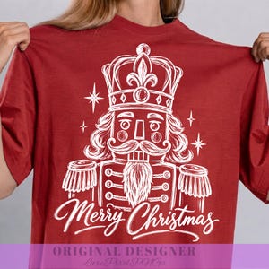 May include: A dark red t-shirt featuring a white line art graphic of a nutcracker wearing a crown. The nutcracker has a moustache and a beard. Below the nutcracker, the text Merry Christmas is written in a white cursive script font.