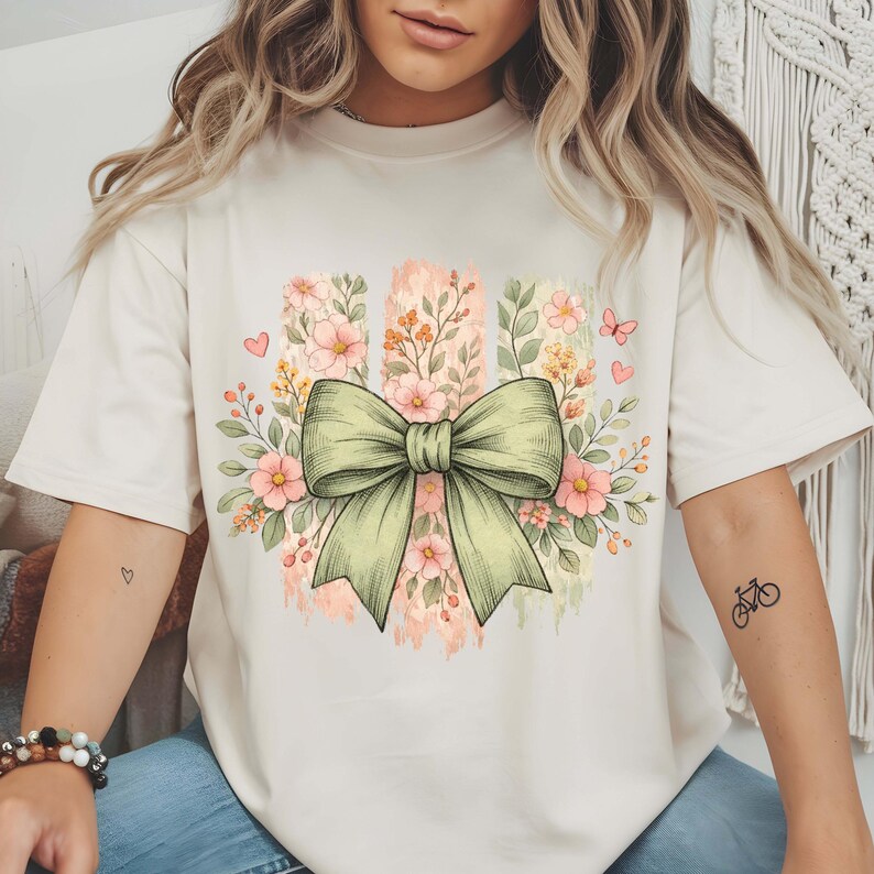 May include: Cream-coloured t-shirt with a large, pale green bow and floral design. The bow is central, surrounded by pink and yellow flowers, green leaves, and small pink hearts. The design is printed on a soft, comfortable tee.