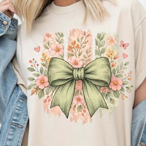 May include: A cream-coloured t-shirt with a large, pale green bow surrounded by pink flowers, leaves, and small hearts. The design includes vertical brushstroke-style stripes in shades of pink and green, creating a floral and feminine aesthetic.