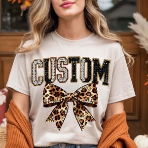Pu&ograve; includere: T-shirt beige con la scritta "CUSTOM" in lettere con strass. Sotto, un fiocco leopardato con decorazioni di strass. Un capo casual per tutti i giorni.