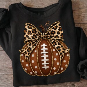 May include: A black long-sleeve sweatshirt featuring a central graphic of a brown pumpkin designed to resemble a football with white laces. The pumpkin is topped with a large leopard print bow and decorated with white polka dots throughout.