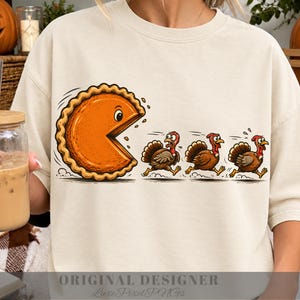 May include: A cream-colored crewneck sweatshirt featuring a graphic print of a large orange pumpkin pie slice with a face chasing three brown turkeys. The pie slice has a scalloped crust edge and is depicted in a cartoon style with motion lines.