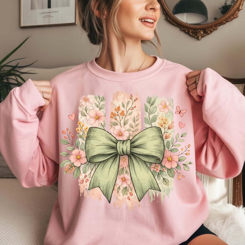 May include: A pale pink sweatshirt with a large green bow and floral design. The design includes pink flowers, green leaves, and a watercolour-style background. The sweatshirt has long sleeves.