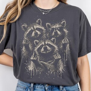 May include: Dark gray t-shirt featuring a graphic of three raccoons giving the middle finger. The raccoons are in a distressed, vintage style with a splatter effect. One raccoon wears glasses. The shirt is a crew neck.