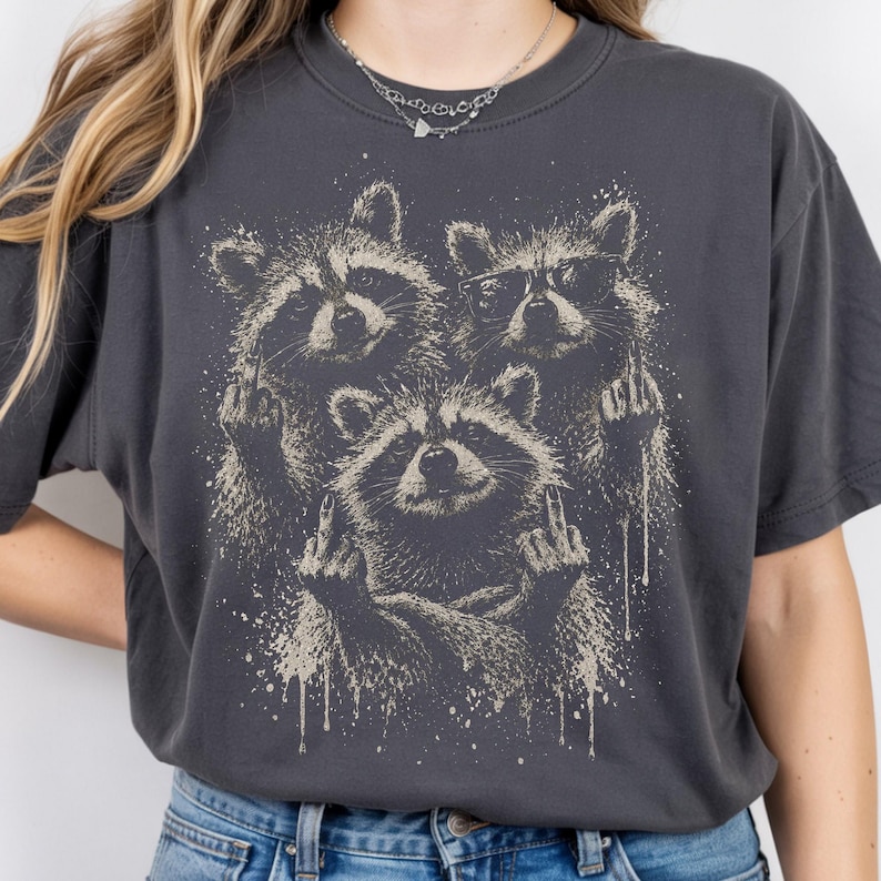 May include: Dark gray t-shirt featuring a graphic of three raccoons giving the middle finger. The raccoons are in a distressed, vintage style with a splatter effect. One raccoon wears glasses. The shirt is a crew neck.