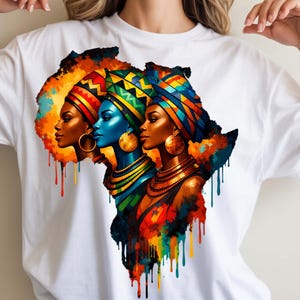 May include: White t-shirt featuring a vibrant graphic of three women's profiles, each adorned with colorful head wraps and jewelry, set against a backdrop resembling the African continent. The artwork uses a rich palette of reds, oranges, blues, and yellows.