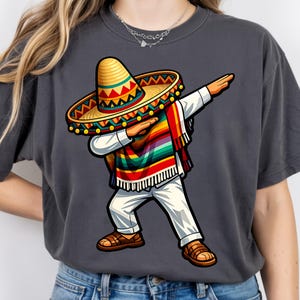 Pode incluir: Camiseta cinza escuro com uma ilustra&ccedil;&atilde;o de desenho animado de uma pessoa fazendo dab, usando um chap&eacute;u, um poncho e cal&ccedil;as brancas. O chap&eacute;u &eacute; amarelo e vermelho, e o poncho &eacute; colorido com arco-&iacute;ris. A pessoa est&aacute; usando sand&aacute;lias marrons.