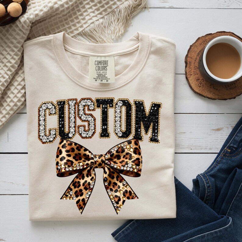 Pu&ograve; includere: T-shirt color crema con la scritta "CUSTOM" in lettere con strass e un fiocco con stampa leopardata. La maglietta &egrave; di Comfort Colors. Una tazza di caff&egrave; e un paio di jeans sono sullo sfondo.