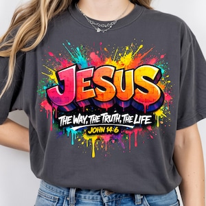 May include: Dark gray t-shirt featuring the word "JESUS" in a graffiti-style design with vibrant pink, orange, and yellow colors. Below the word, the text reads "THE WAY, THE TRUTH, THE LIFE" and "JOHN 14:6". The design includes colorful paint splatters.