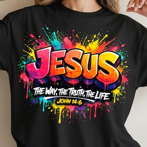 May include: Black t-shirt featuring the word "JESUS" in a graffiti-style design with vibrant pink, orange, and yellow lettering. The shirt also displays the text "THE WAY, THE TRUTH, THE LIFE" and "JOHN 14:6" against a colorful paint splatter background.