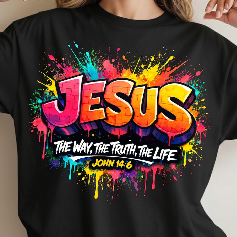 May include: Black t-shirt featuring the word "JESUS" in a graffiti-style design with vibrant pink, orange, and yellow lettering. The shirt also displays the text "THE WAY, THE TRUTH, THE LIFE" and "JOHN 14:6" against a colorful paint splatter background.