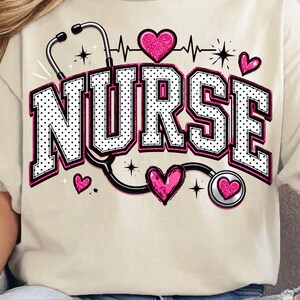 Puede incluir: Una camiseta de color crema con un estampado gr&aacute;fico que muestra la palabra NURSE en letras blancas grandes con puntos negros y bordes rosas. Un estetoscopio negro rodea el texto, decorado con corazones rosas brillantes y estrellas negras.