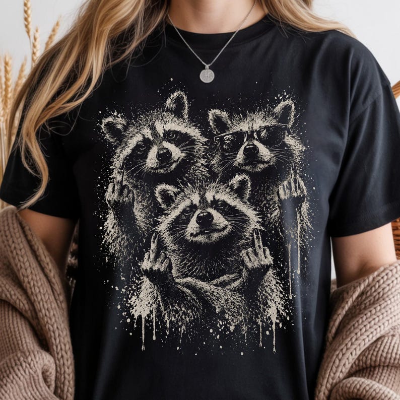 May include: Black t-shirt featuring a graphic of three raccoons. The raccoons are depicted in a black and white illustration, each with a hand gesture. One raccoon wears sunglasses. The design has a distressed, splattered effect.