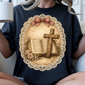 May include: Dark navy blue t-shirt with a sepia-toned graphic of an open book, a wooden cross, and roses within a lace-edged oval. A pink bow is at the top. The design has a vintage, religious theme.