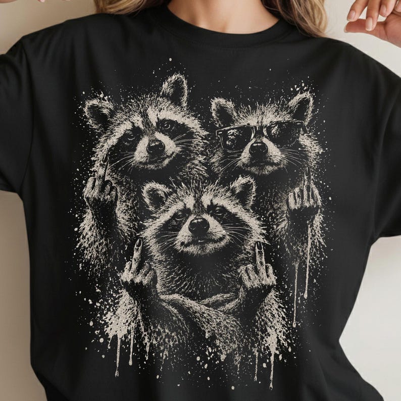 May include: Black t-shirt featuring a graphic of three raccoons. The raccoons are depicted in a detailed, grayscale illustration, with two raccoons giving the middle finger. One raccoon wears sunglasses. The design includes white paint splatter.