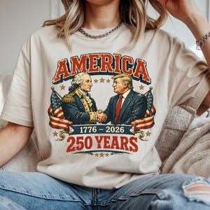 May include: A beige t-shirt featuring a graphic with the word "AMERICA" in red and white, above an illustration of two men shaking hands. The graphic includes the text "1776 - 2026" and "250 YEARS" in red.