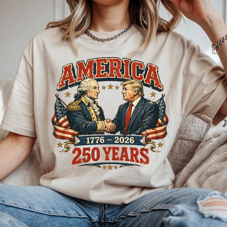 May include: A beige t-shirt featuring a graphic with the word "AMERICA" in red and white, above an illustration of two men shaking hands. The graphic includes the text "1776 - 2026" and "250 YEARS" in red.