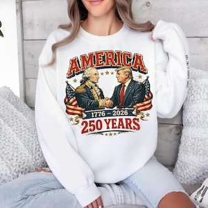 May include: White sweatshirt with a graphic featuring the word "AMERICA" in red, with an illustration of two men shaking hands, American flags, and the text "1776-2026 250 YEARS".