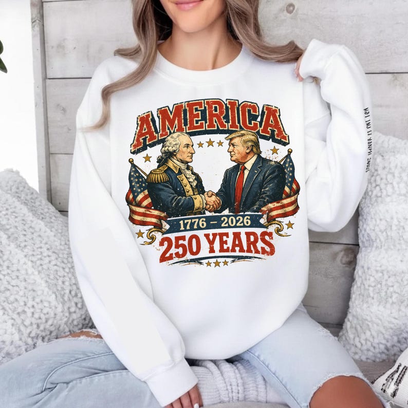 May include: White sweatshirt with a graphic featuring the word "AMERICA" in red, with an illustration of two men shaking hands, American flags, and the text "1776-2026 250 YEARS".