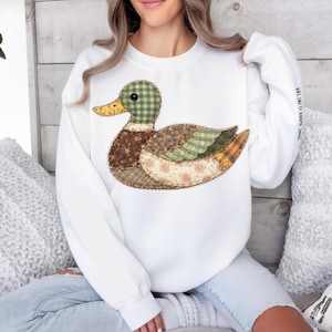 May include: White sweatshirt featuring a patchwork duck design. The duck has a green checkered head, brown body, and wings with floral and plaid patterns. The sleeve has the text "STRONG, HANNAH, IS MY NAME".