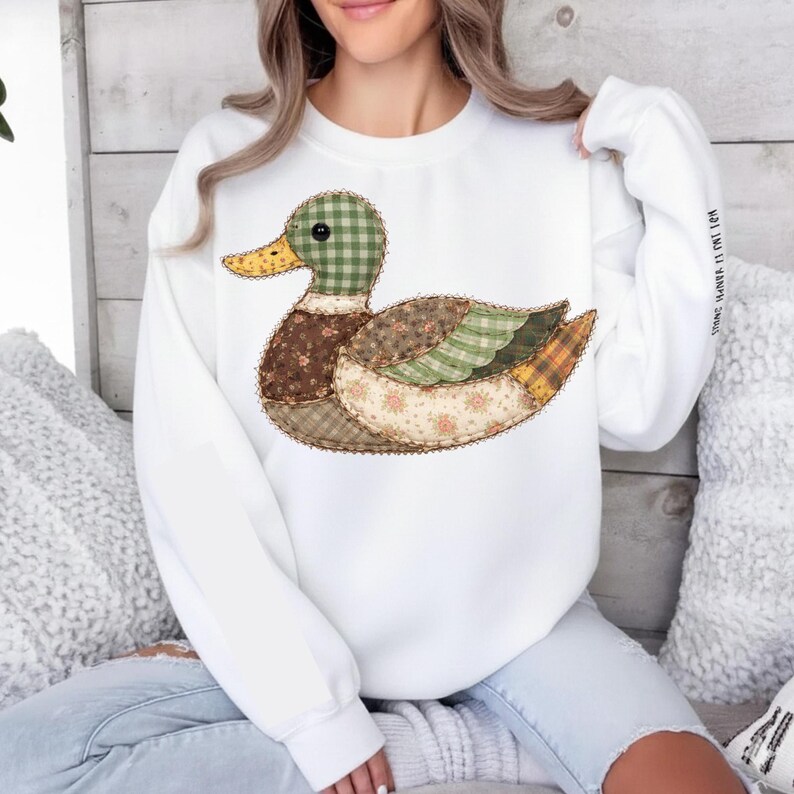 May include: White sweatshirt featuring a patchwork duck design. The duck has a green checkered head, brown body, and wings with floral and plaid patterns. The sleeve has the text "STRONG, HANNAH, IS MY NAME".