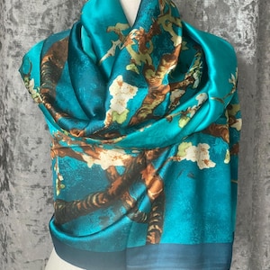 May include: A teal silk scarf featuring a print of a tree branch with white flowers. The scarf is draped over a mannequin, highlighting its flowing fabric and design. The scarf has a dark teal border.